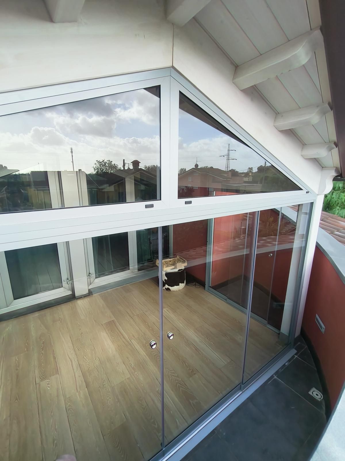 Balcone con pareti in vetro, finestra superiore triangolare e pavimento in legno chiaro, che riflette il cielo.