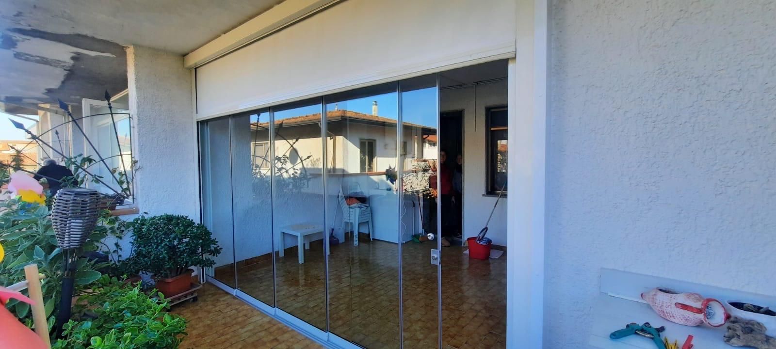 Balcone con porte scorrevoli in vetro che riflettono l'esterno. Piante e mobili a vista. Pareti bianche. Giornata di sole.