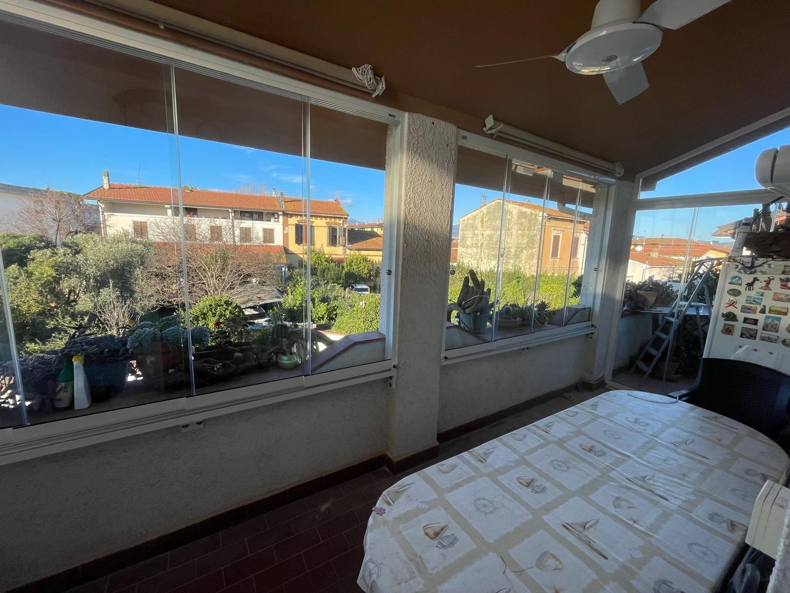 Veranda chiusa e soleggiata con vetrate che si affacciano sul verde e sugli edifici, tavolo apparecchiato.