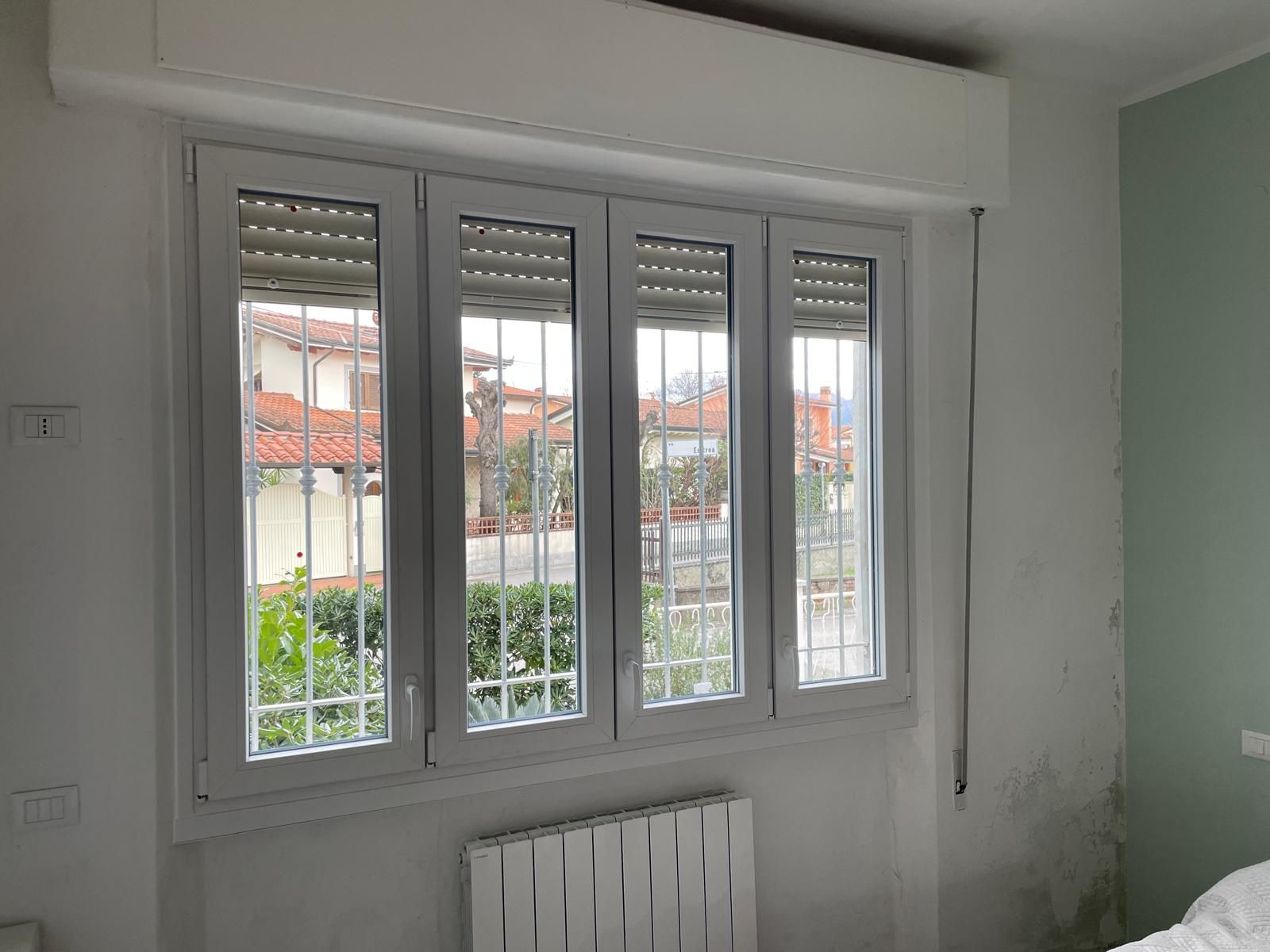 Finestra a quattro vetri con cornici bianche, tende avvolgibili, inferriate di sicurezza e vista sugli edifici e sul verde.