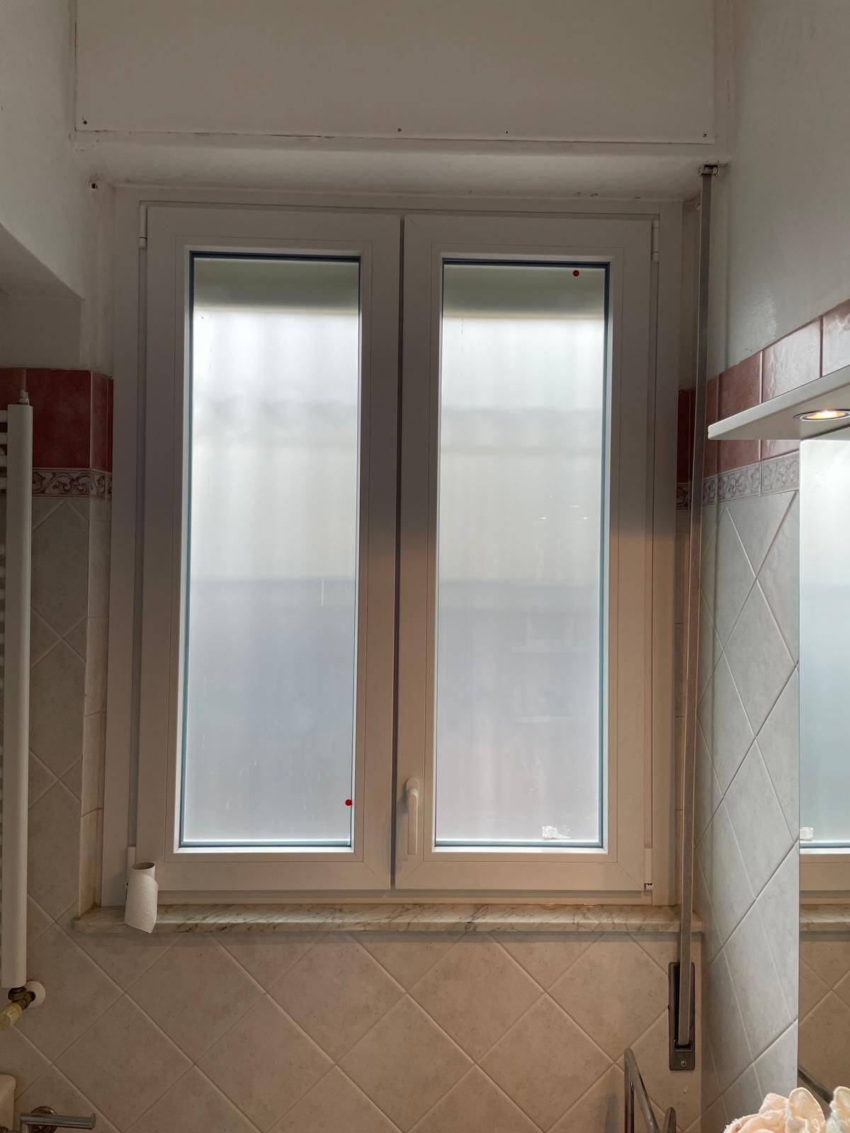 Finestra a due ante con cornice bianca e vetro smerigliato in un ambiente da bagno, sopra una superficie piastrellata.