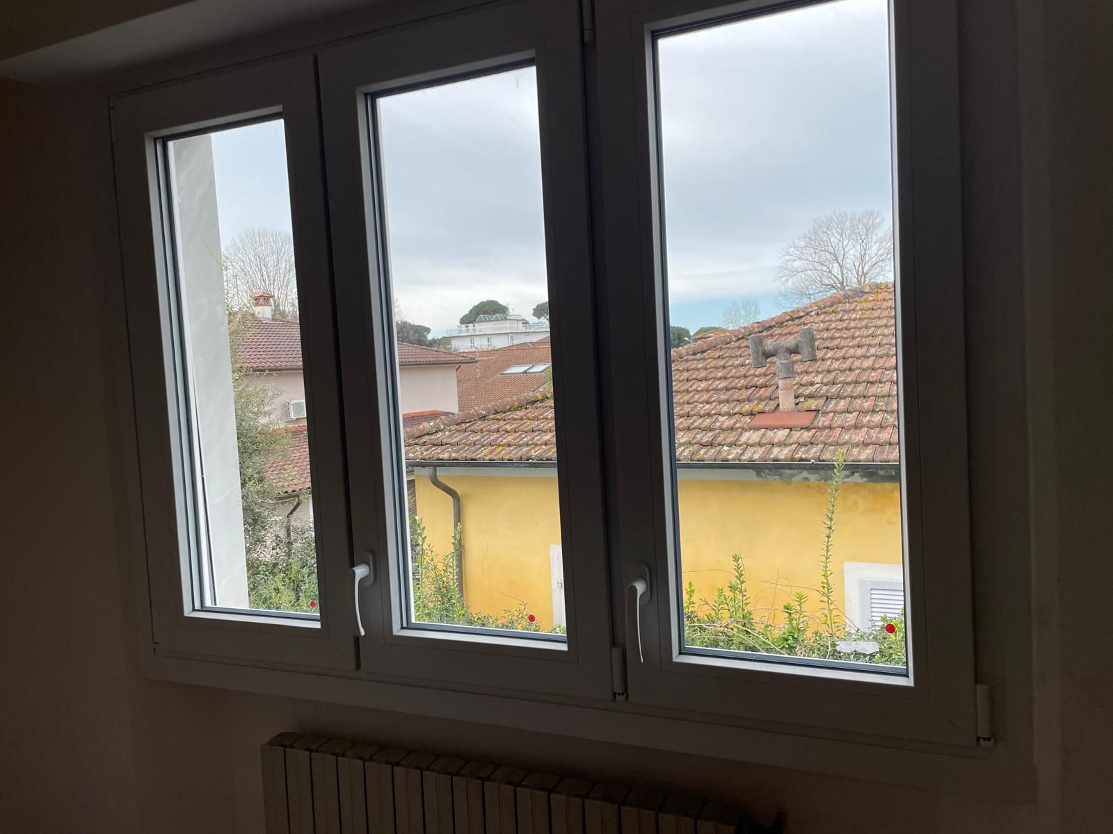 Finestra a tre vetri con vista sui tetti, un edificio giallo e un cielo nuvoloso. Sotto la finestra c'è un termosifone.