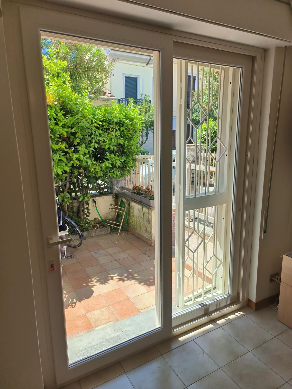 Le portefinestre con cornice bianca si aprono su un patio in mattoni con vegetazione lussureggiante e una bicicletta.