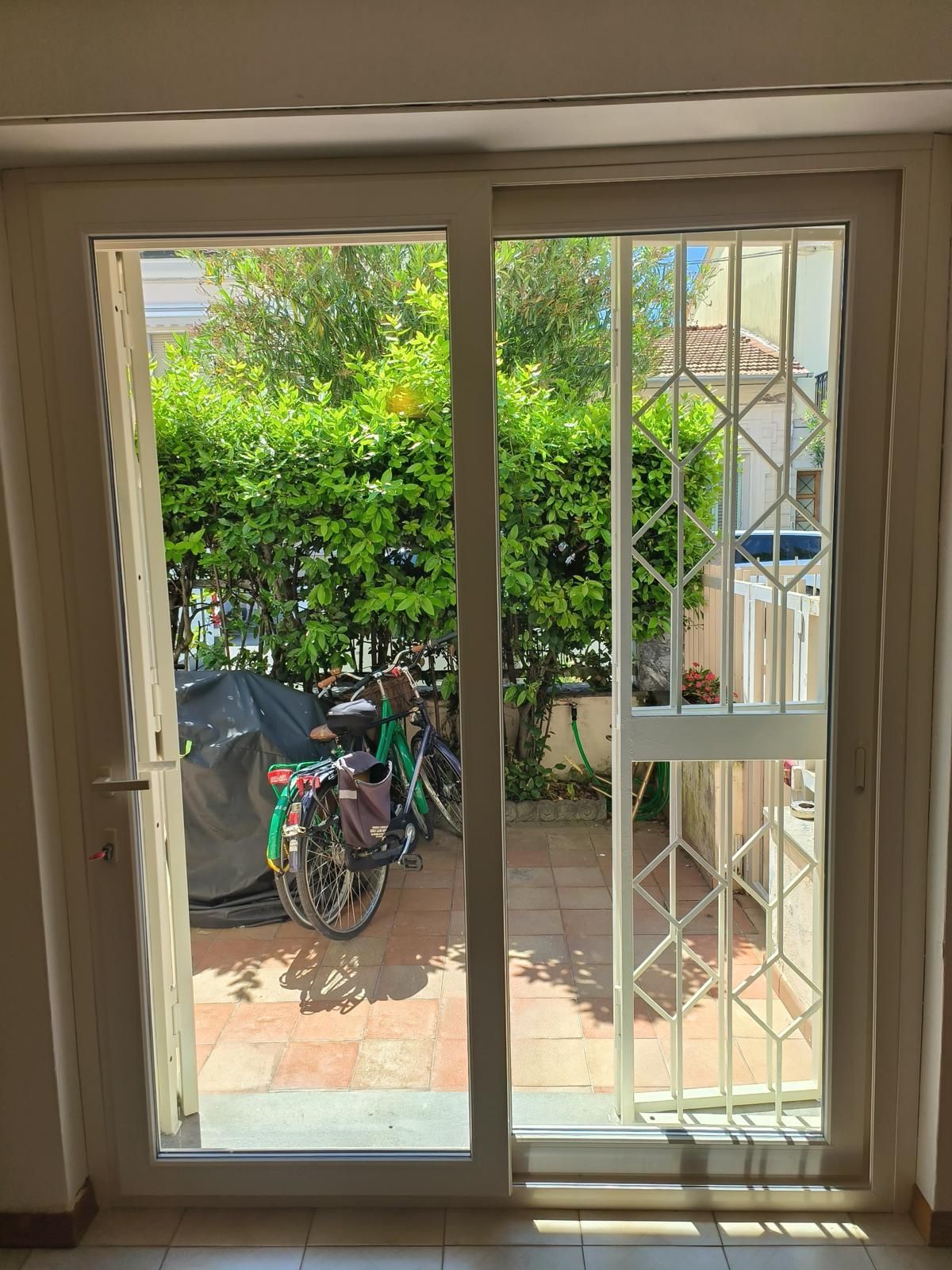Porta scorrevole in vetro con cancelletto di sicurezza bianco che si apre su un patio con biciclette e verde. 