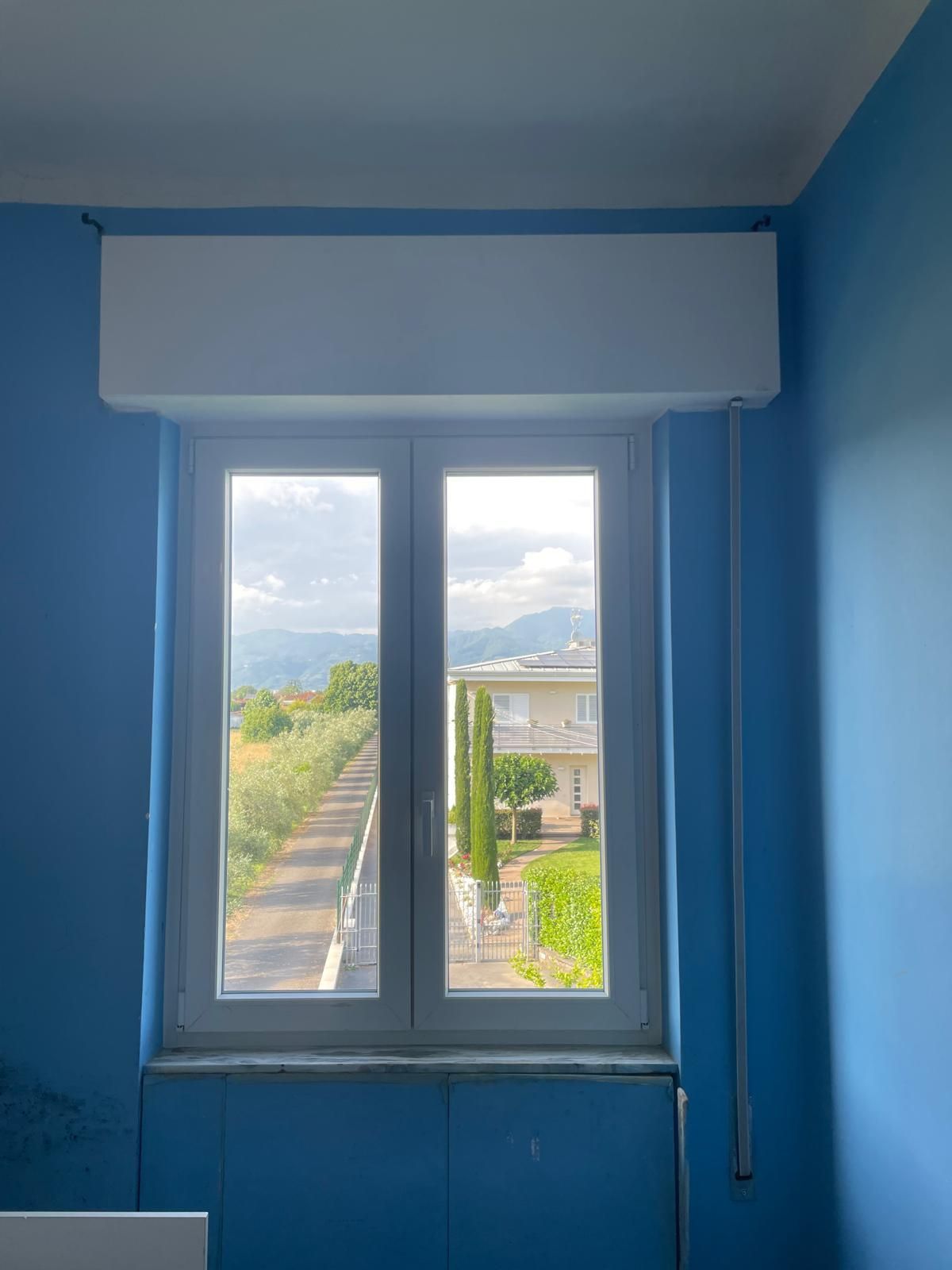 Finestra con cornice bianca, pareti blu, vista su una strada e degli edifici e una scatola bianca sopra la finestra.