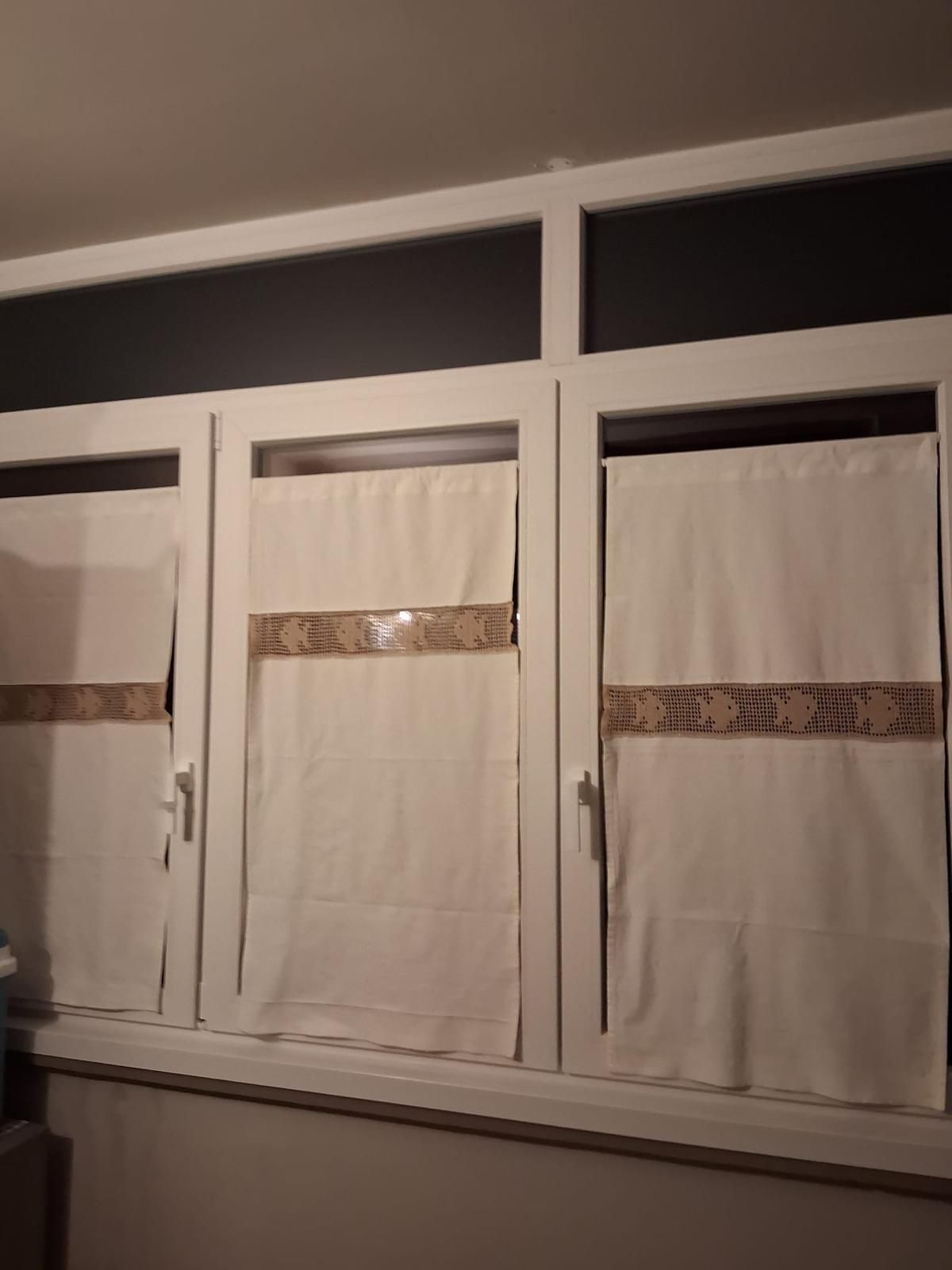 Tre finestre con cornice bianca e tende chiare fissate con nastro adesivo, addossate a una parete scura.