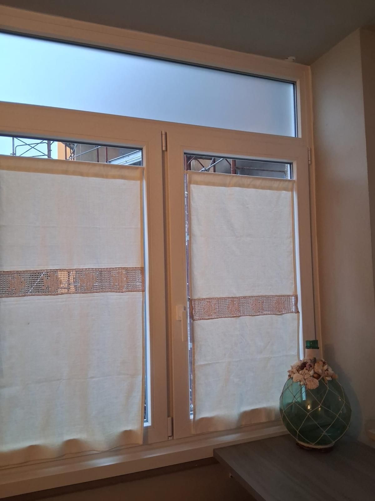 Finestra con cornice bianca, vetro superiore satinato e due tende avvolgibili. Un vaso è appoggiato sul davanzale.