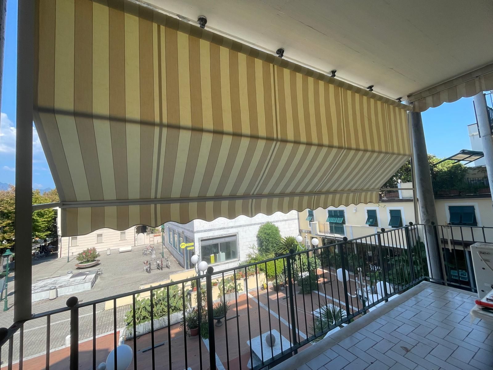 Una tenda a strisce ombreggia un balcone che si affaccia su un cortile con alberi ed edifici.