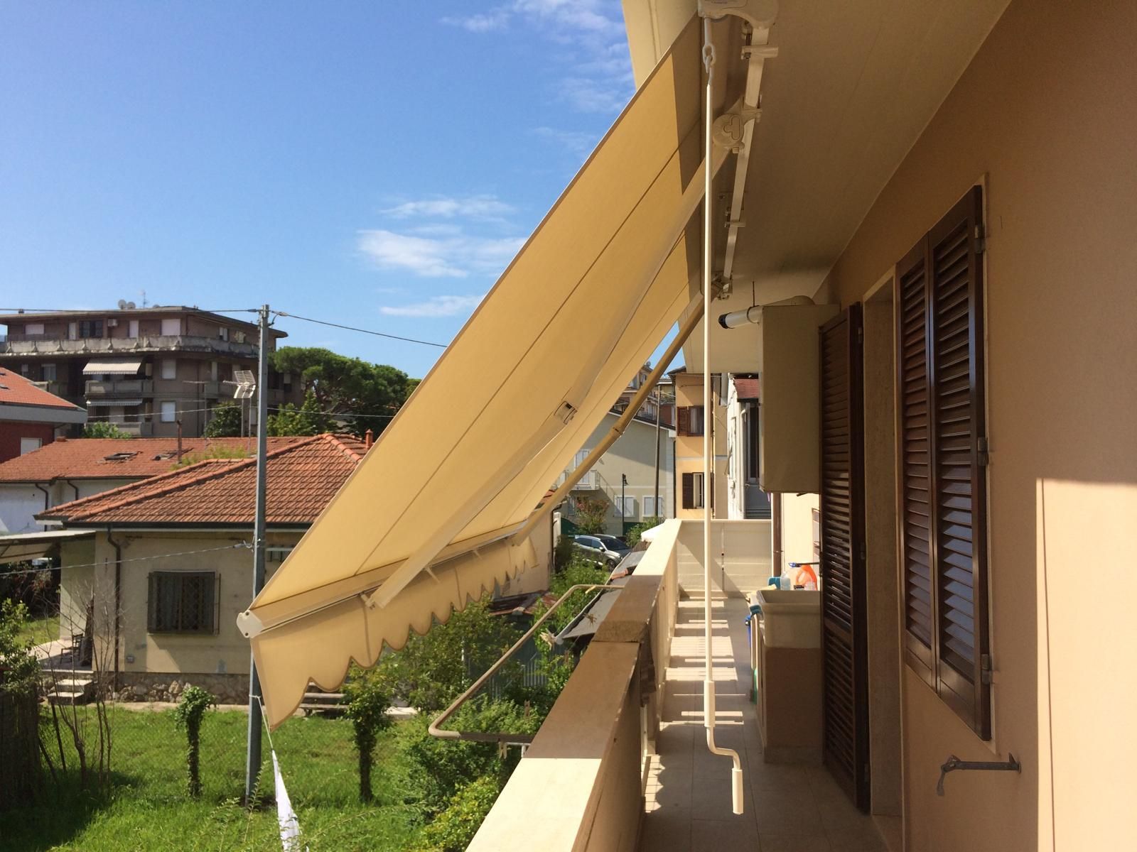 Tenda da sole beige che si estende su un balcone, accanto a un edificio con persiane marroni. Cielo azzurro sullo sfondo.