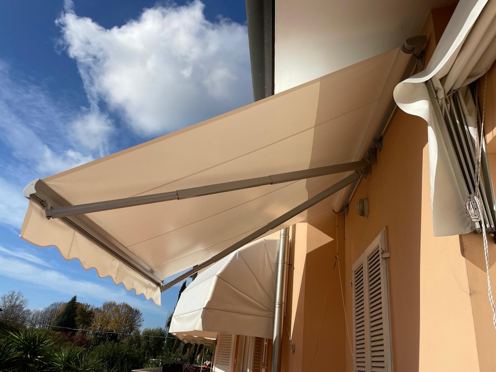 Tenda da sole retrattile beige stesa su un edificio color pesca, contro un cielo parzialmente nuvoloso.