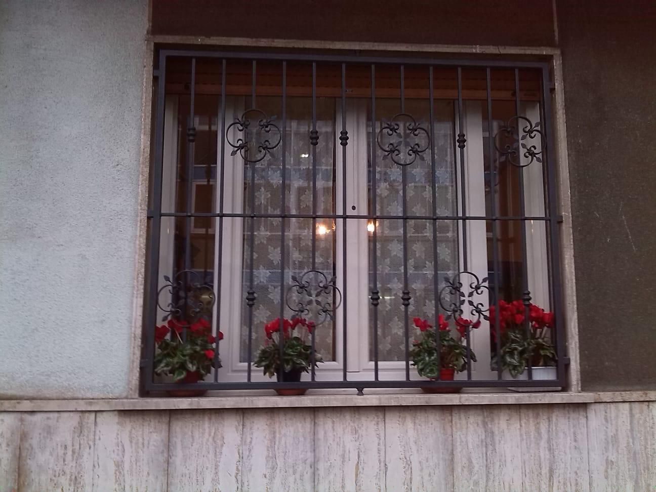 Finestra con inferriate decorative, fiori rossi in vaso e tende bianche.