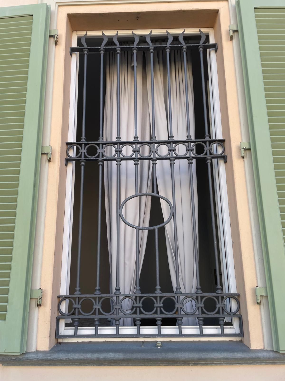 Finestra con inferriate di sicurezza in ferro battuto grigio, tende leggere e persiane verdi.