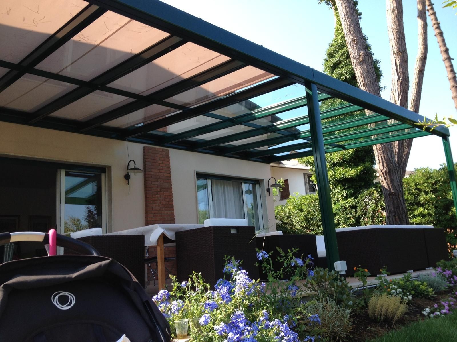 Copertura per patio con cornice verde sopra una terrazza con piante e un oggetto scuro in primo piano, giornata di sole.