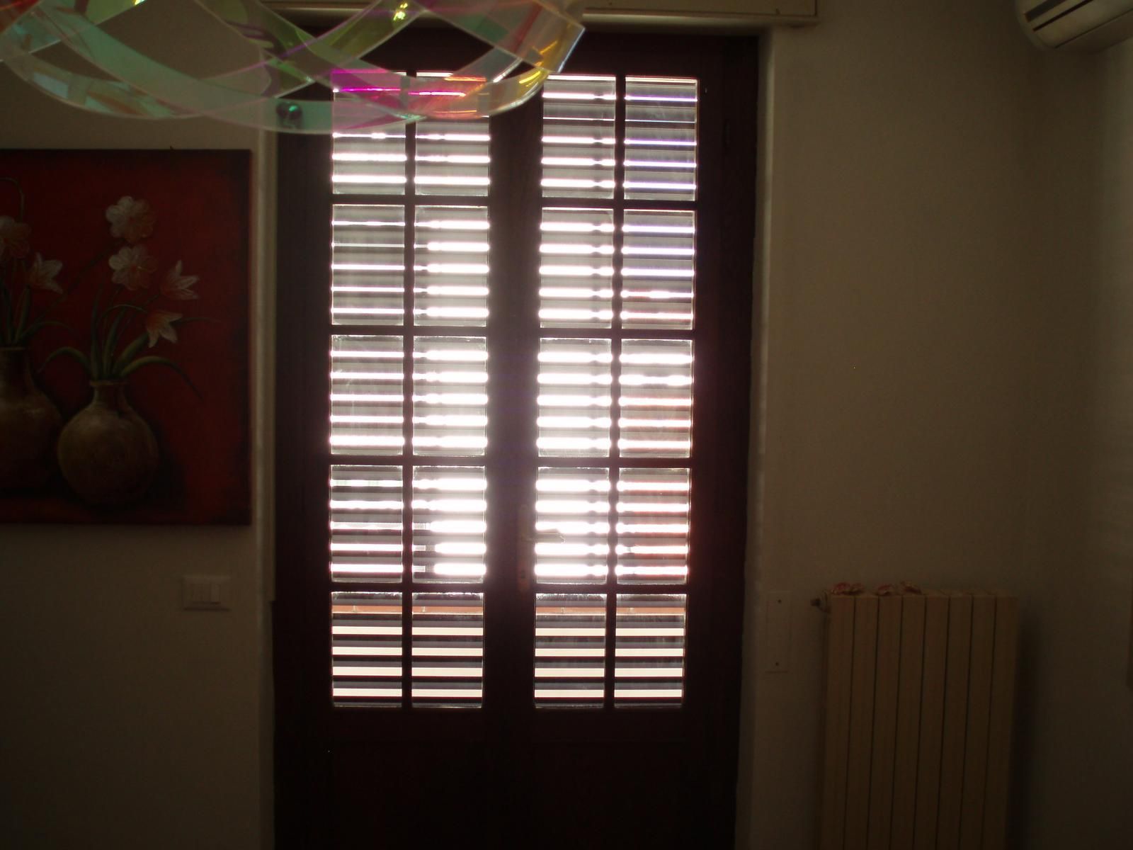 Porta marrone con persiane orizzontali, luce che filtra attraverso. Dipinto di vasi sulla sinistra.