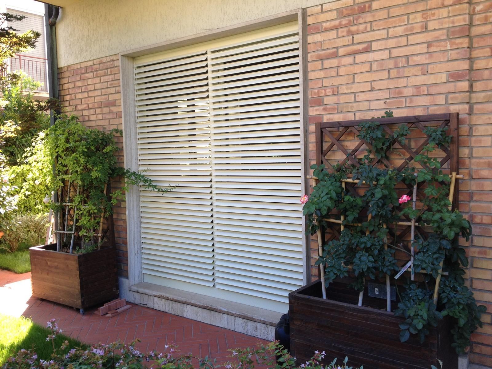 Finestra con persiane chiare chiuse, affiancata da fioriere in legno scuro con piante verdi contro un muro di mattoni.