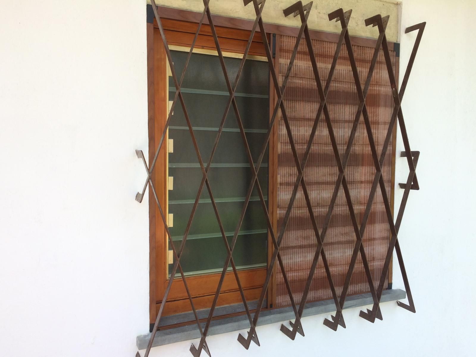 Finestra con grata di sicurezza marrone a fisarmonica, parzialmente aperta, contro una parete esterna bianca.