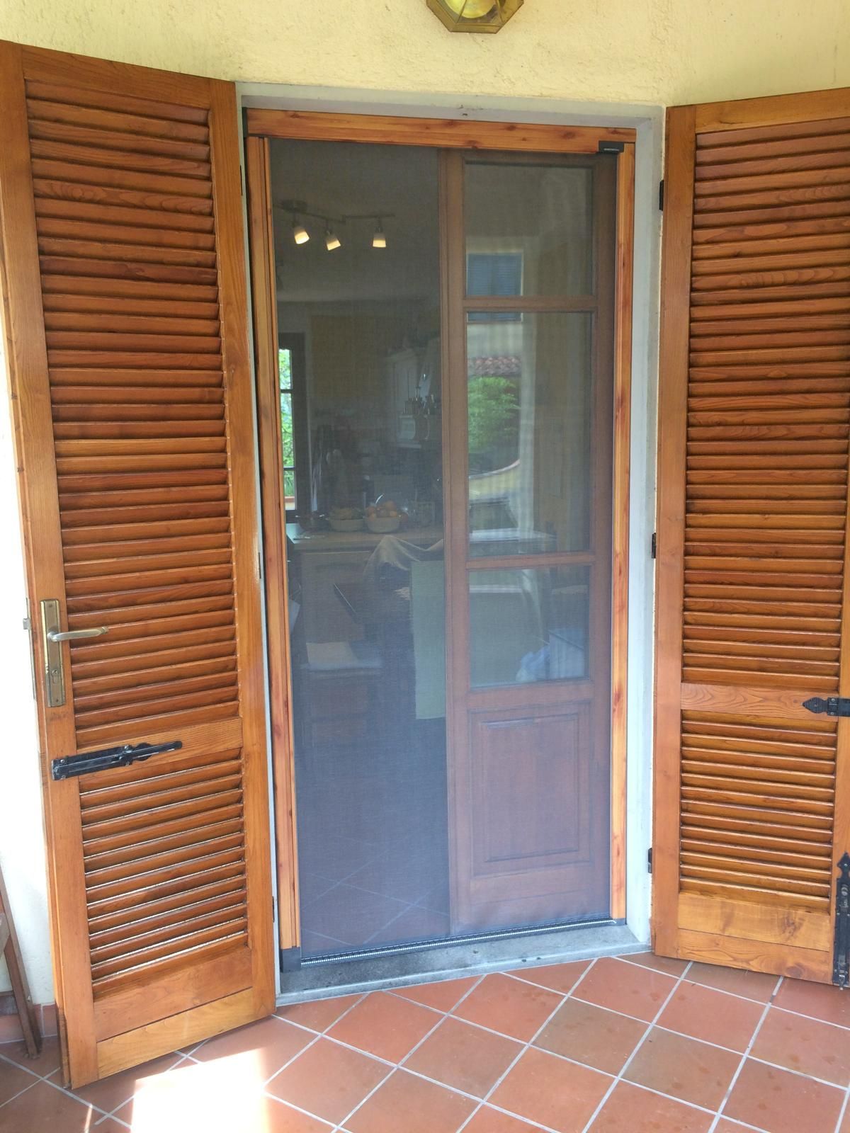 Porte aperte in legno con persiane che fiancheggiano una porta a zanzariera; pavimento in piastrelle di terracotta.
