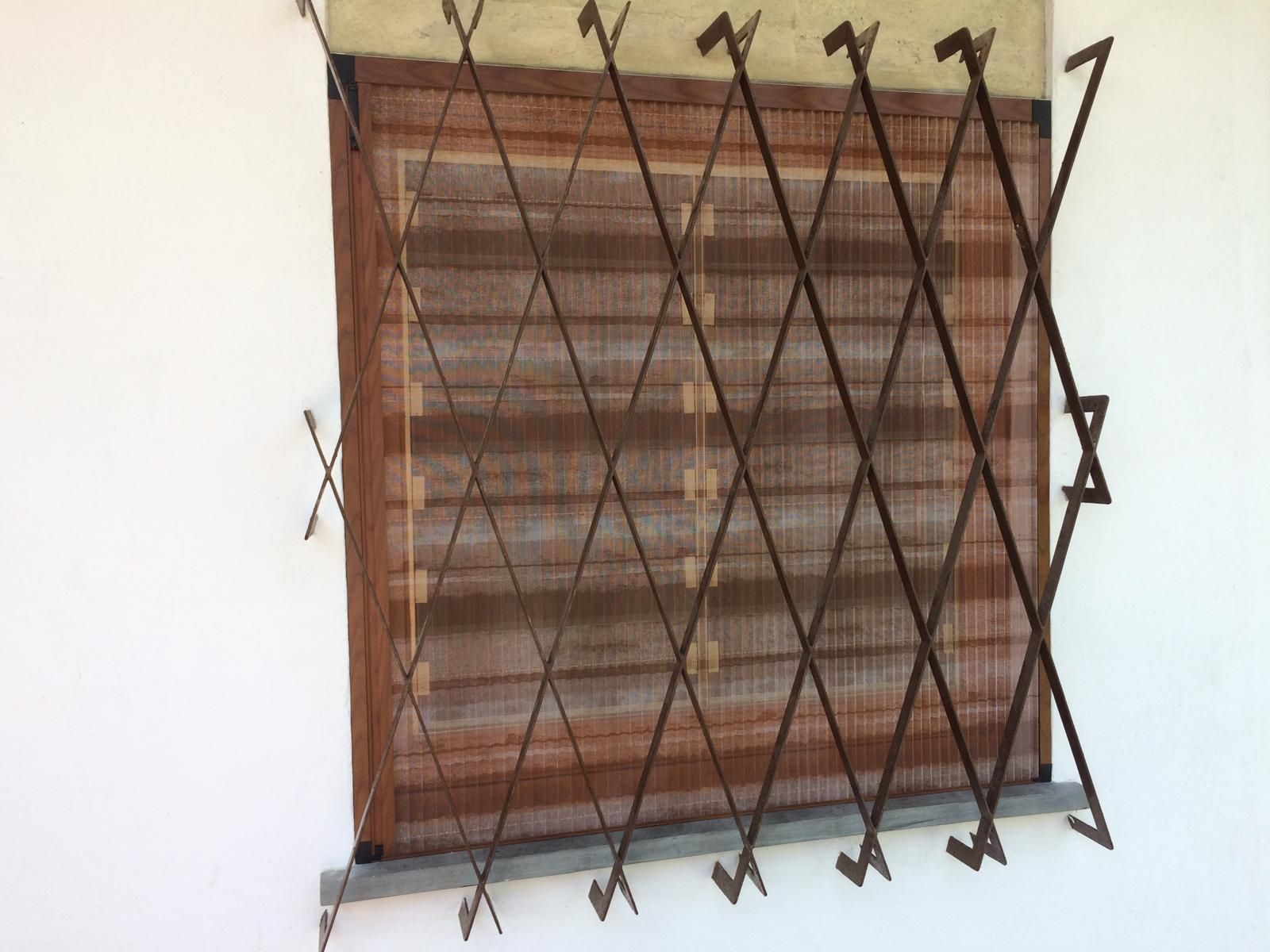 Finestra con una griglia di sicurezza marrone a fisarmonica e una zanzariera marrone contro una parete bianca.