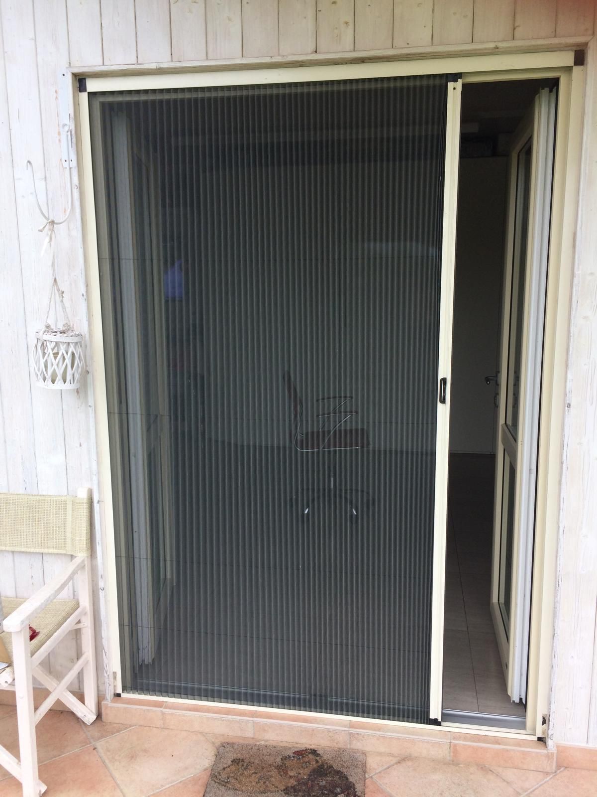 Porta scorrevole con telaio beige, aperta verso l'interno, rete verticale scura. Portico, sedia a vista.