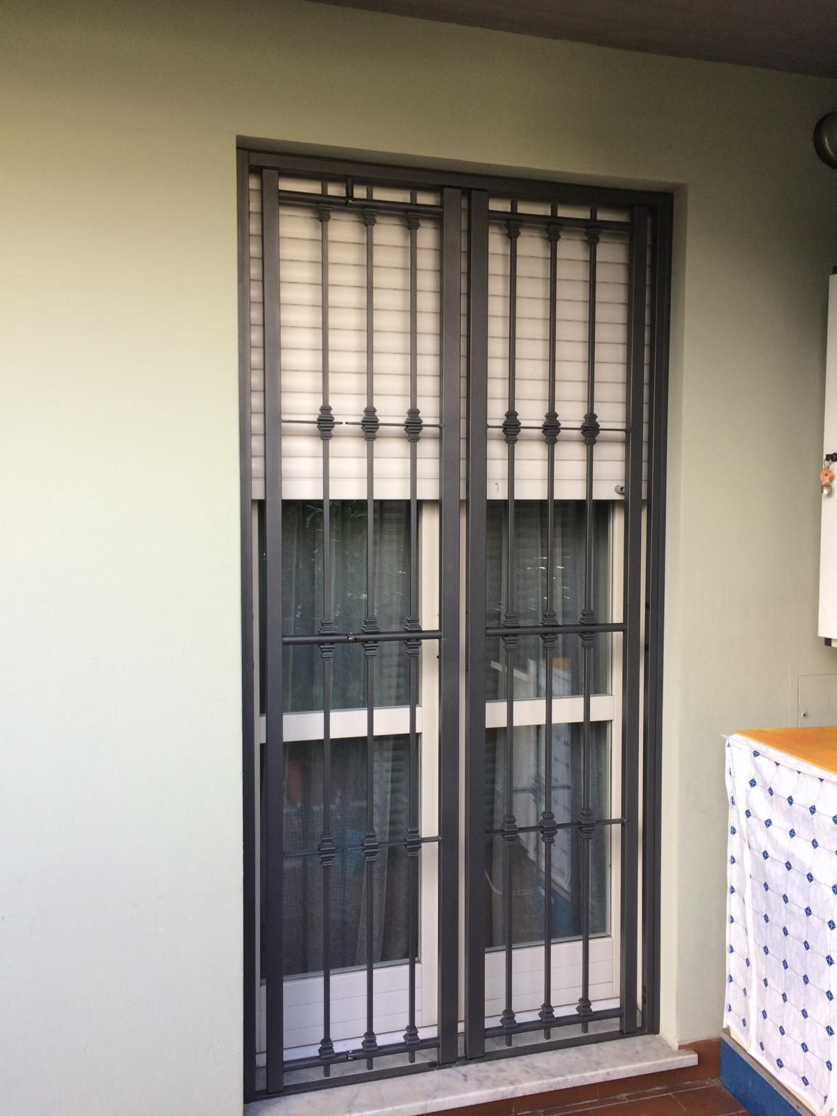 Barre di sicurezza metalliche che coprono una porta doppia bianca e delle persiane, contro una parete verde chiaro.