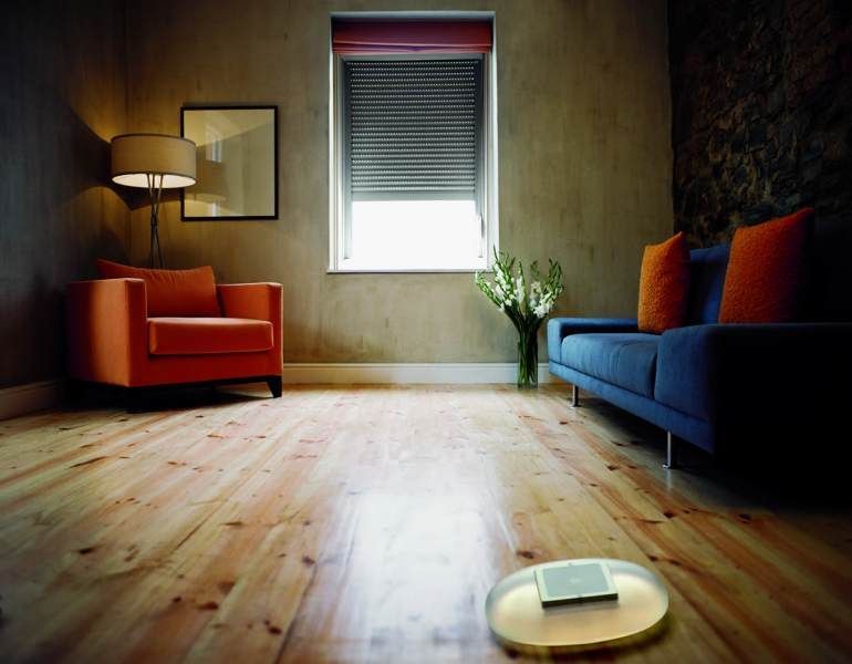 Soggiorno con pavimento in legno, sedia arancione, divano blu, finestra con paralume, lampada e fiori.