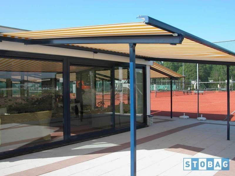 Tenda a strisce gialle e nere su un edificio con pareti in vetro, sostenuta da pali di metallo blu, su un patio in mattoni.