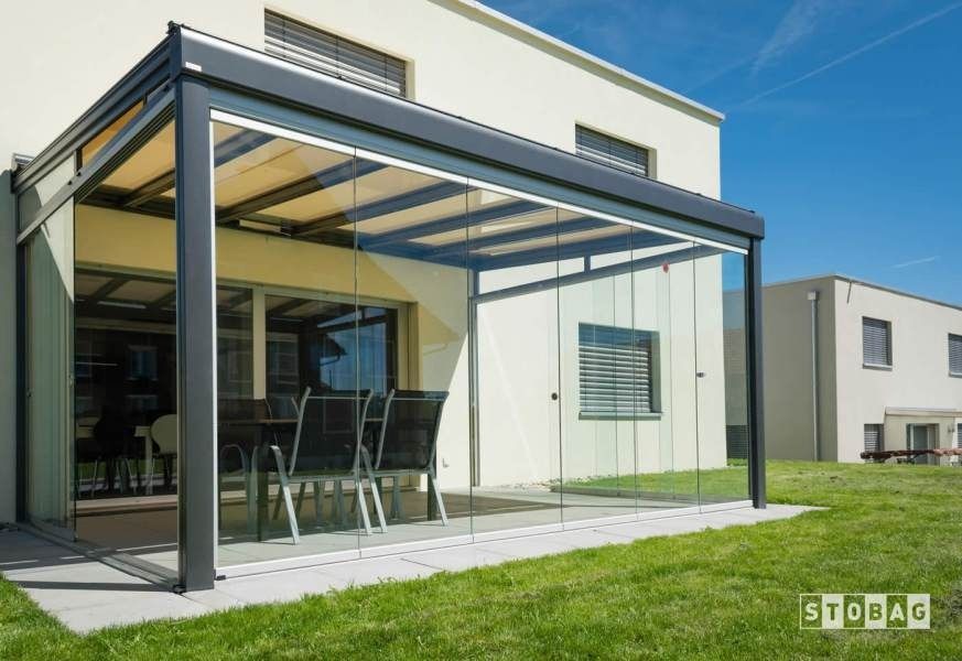 Moderna recinzione per patio con pareti in vetro, struttura nera e tetto retrattile; tavolo e sedie all'interno.
