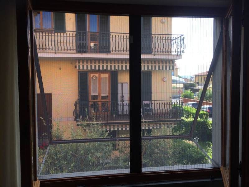 Finestra aperta con zanzariera che si affaccia su un condominio, sul verde e sui veicoli.