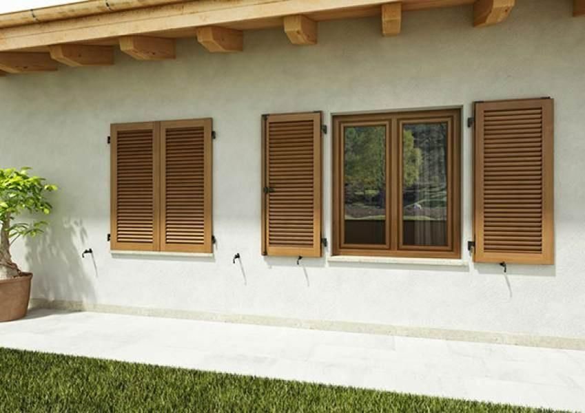 Esterno di una casa bianca con tre finestre e persiane in legno marrone; pianta in vaso sulla sinistra, erba verde sotto.