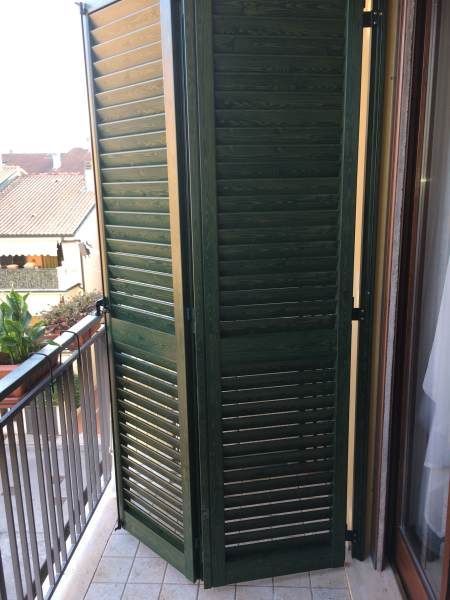 Persiane verdi su un balcone, parzialmente aperte. Sono visibili una ringhiera metallica e un edificio.