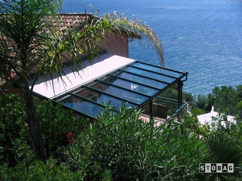 Terrazza con tetto in vetro e vista mare, circondata dal verde. Logo STOBAG ben visibile.