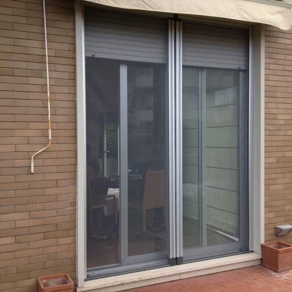 Porta scorrevole in vetro con zanzariera e persiane grigie chiuse, inserita in un muro di mattoni.
