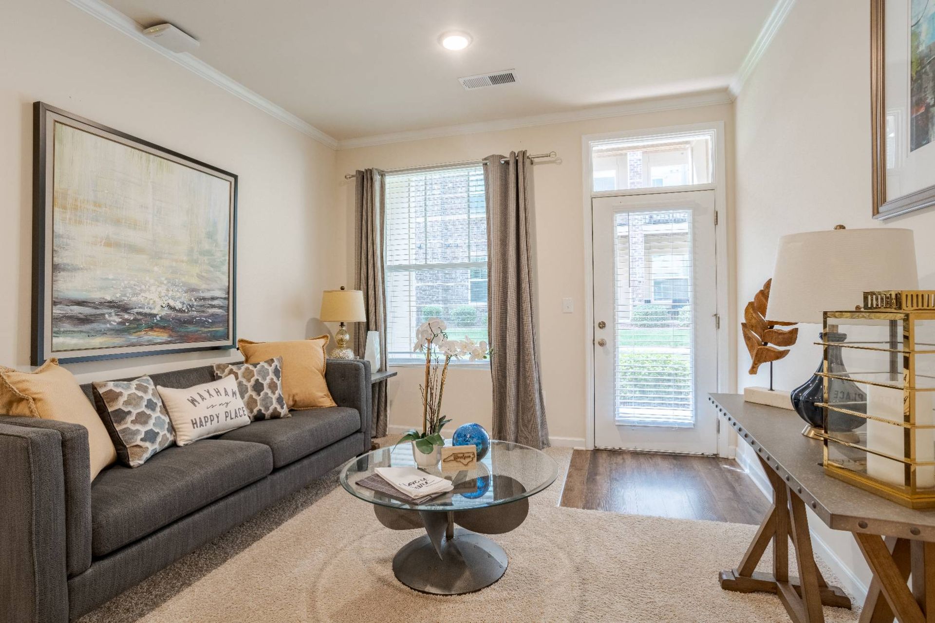 Living room in an apartment with a gray sofa, patterned pillows, glass coffee table, and a door to outdoors. 