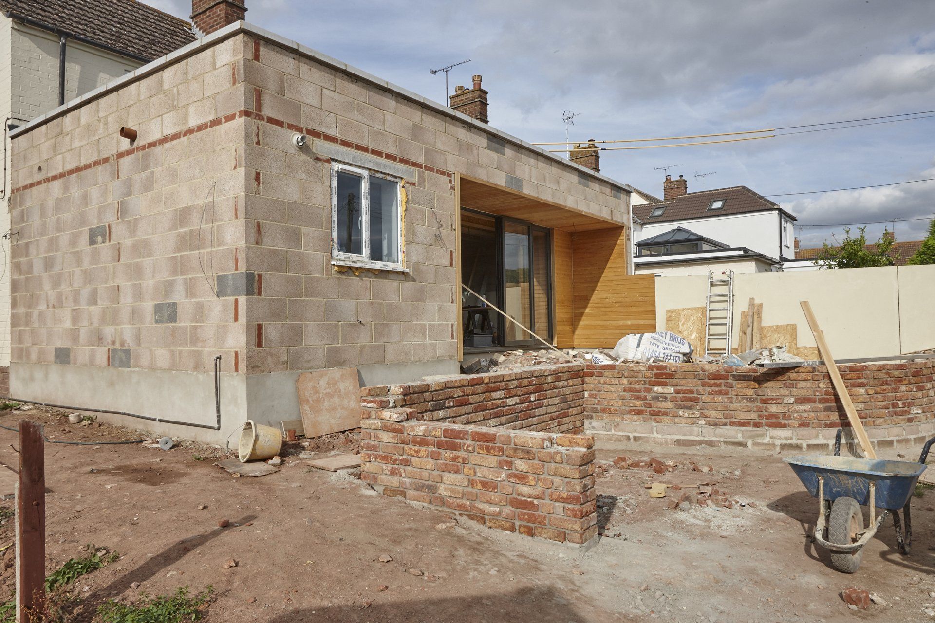 A house is being built with bricks and a wheelbarrow in front of it.