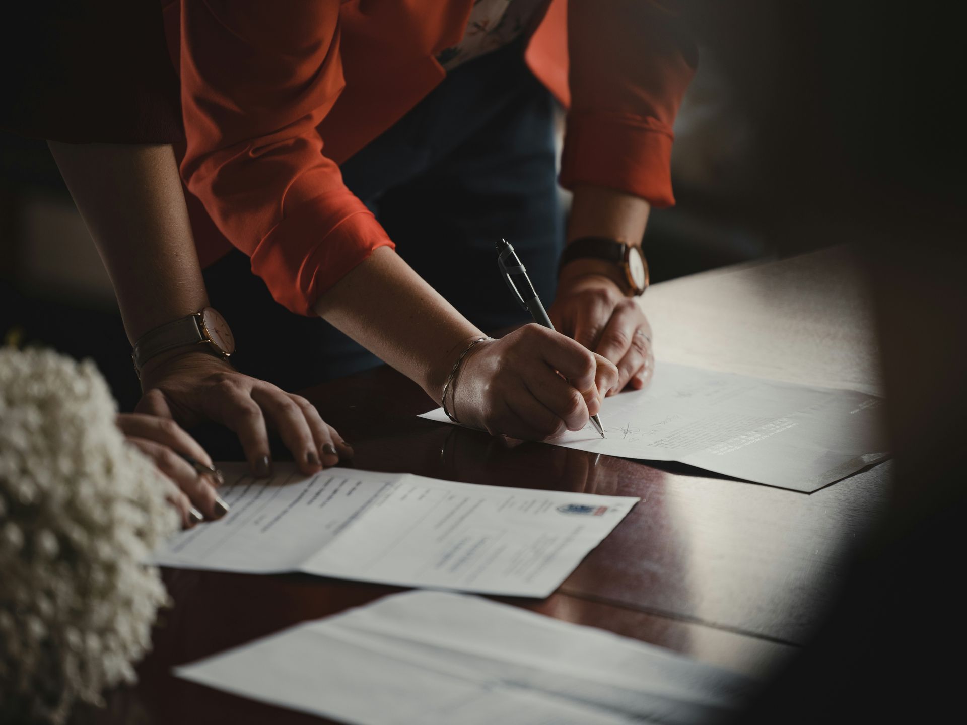 Hands signing documents on a dark wooden table; one person wears a red shirt.