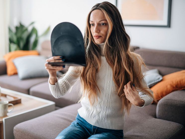 A woman is sitting on a couch looking at her hair in a mirror.