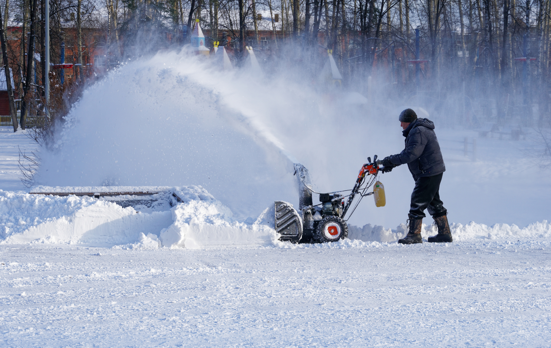 A person uses a snowblower to clear snow from a paved area. White snow is blown upwards in a large arc.