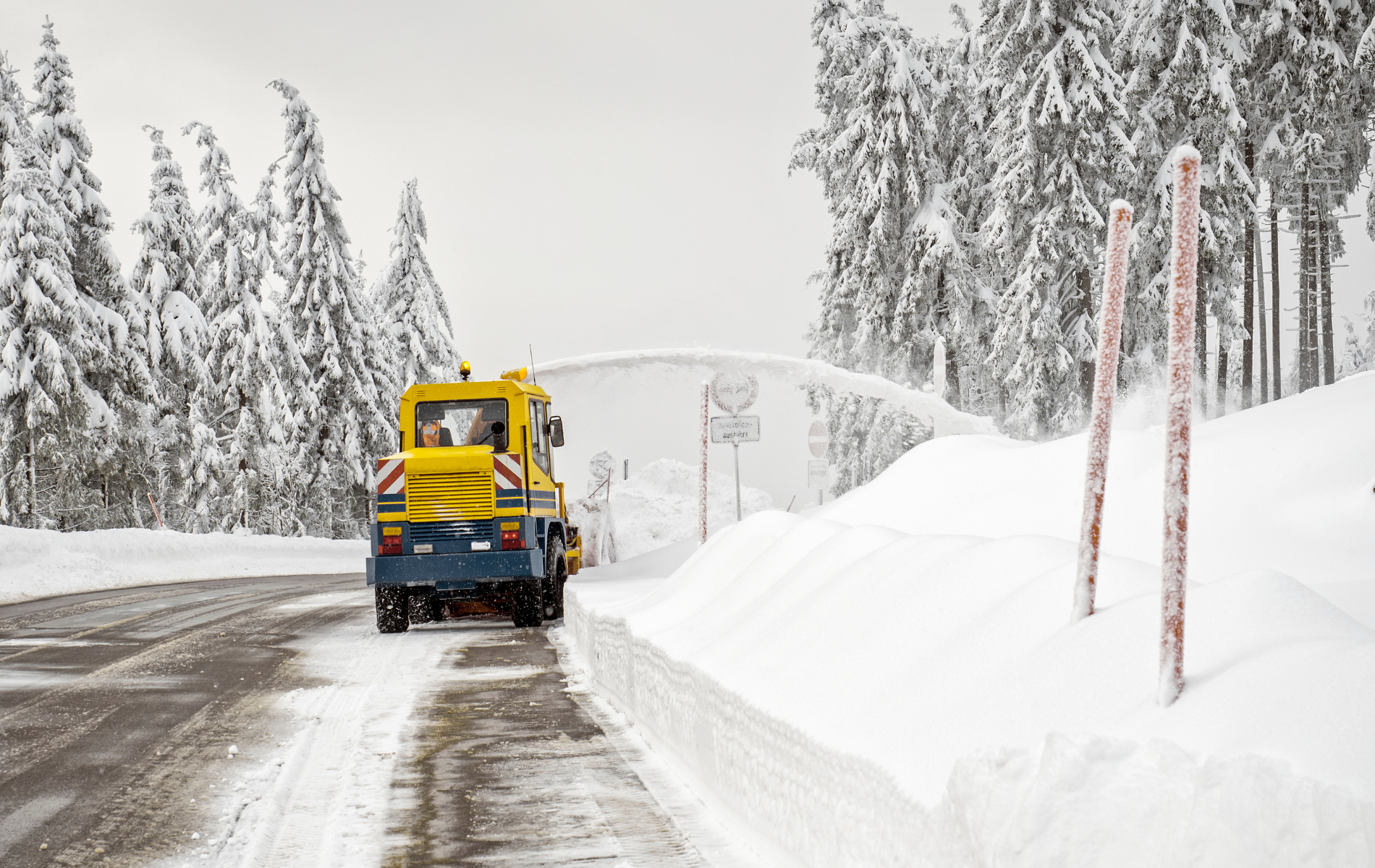 A yellow snowplow clearing a snow-covered road in a forest, pushing snow to the side.