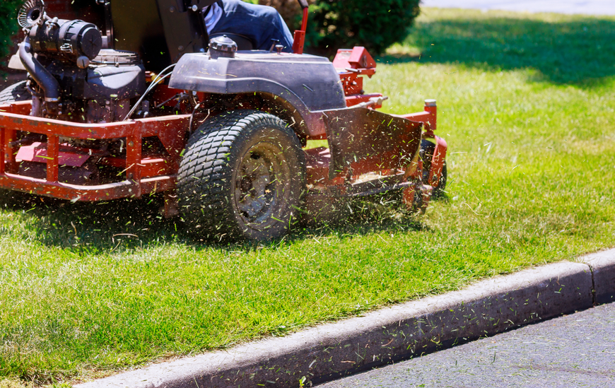 A red zero-turn lawnmower cuts grass next to a curb, with grass clippings flying.