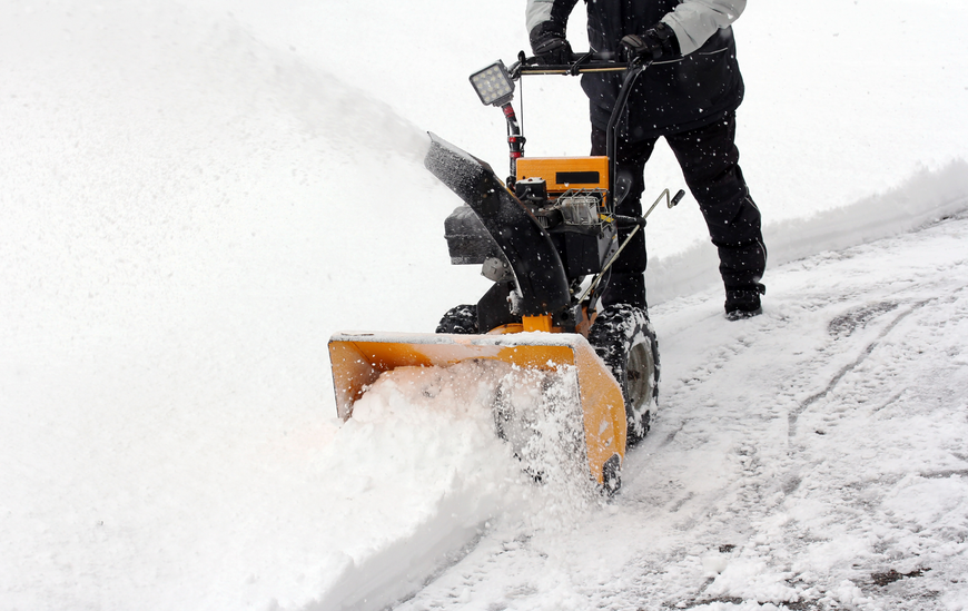 A person using a yellow snowblower to clear snow from a white-covered driveway.