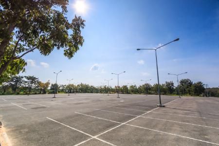 Empty asphalt parking lot under a bright blue sky with sparse trees and lampposts.