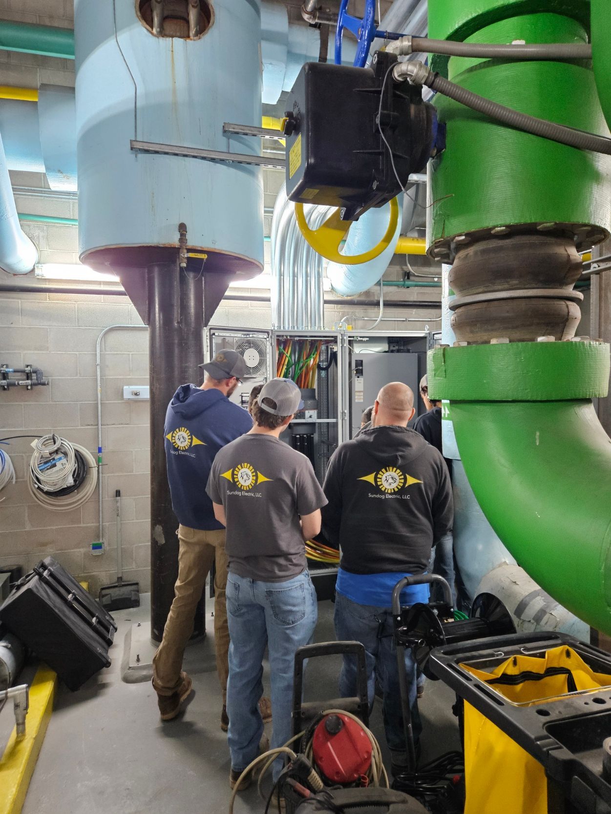 Three workers in a utility room, looking at a control panel; large pipes and equipment surround them.