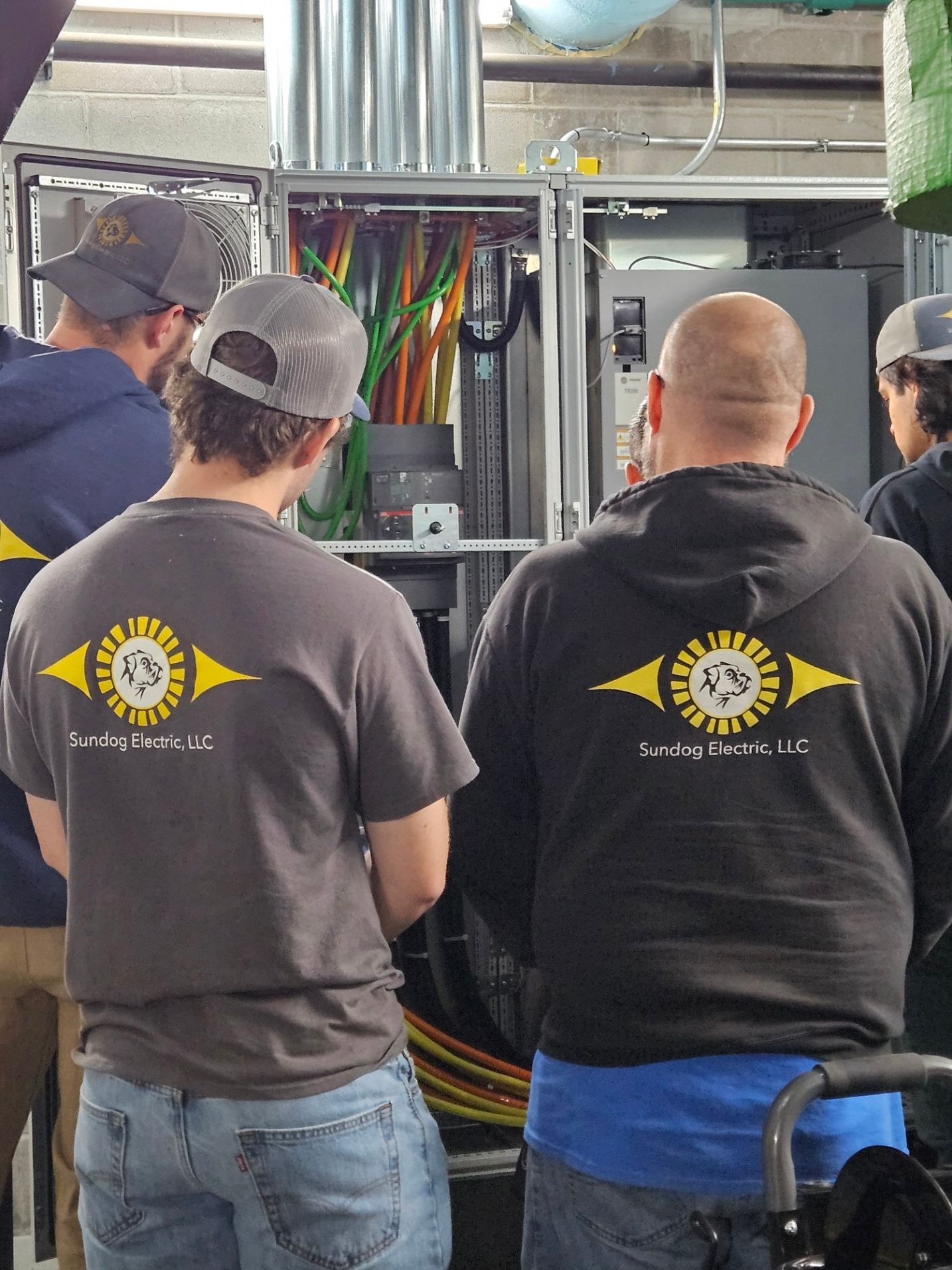 Group of electricians working on an electrical panel, wearing matching t-shirts, in an industrial setting.
