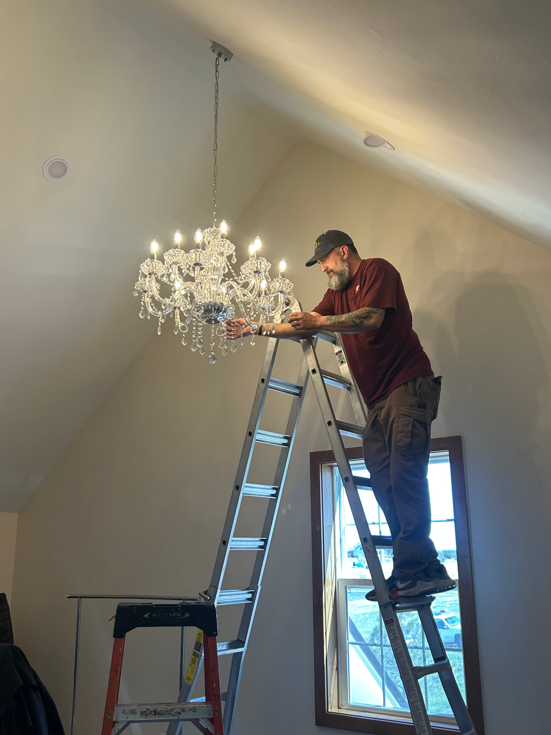 Man on a ladder installs a chandelier in a room with a slanted ceiling and a window.