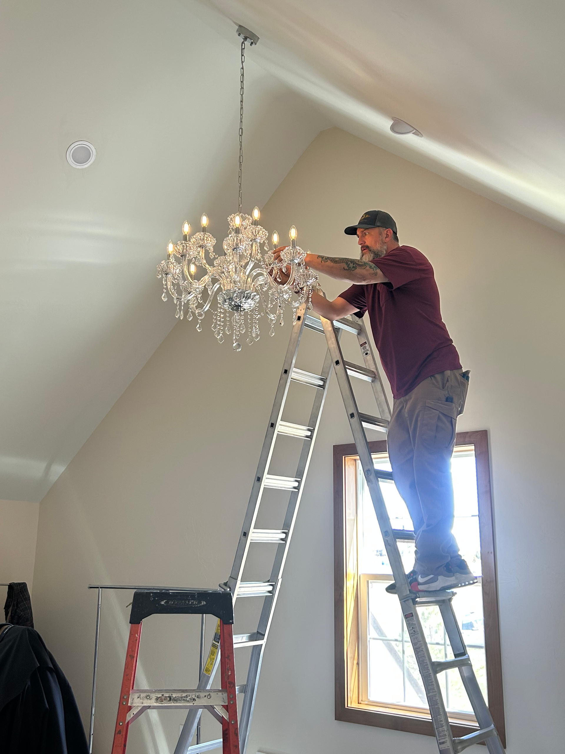 Man on ladder adjusting a crystal chandelier in a room with a sloped ceiling.