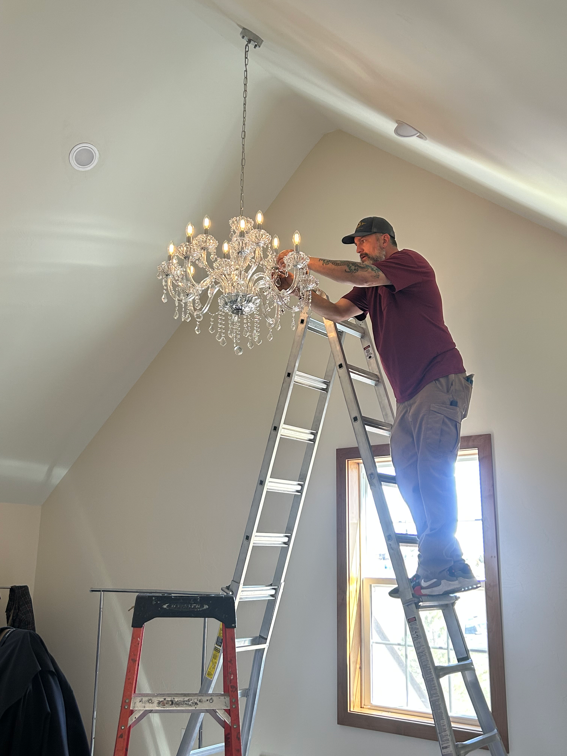 Man on ladder installing a crystal chandelier in a light-colored room with angled ceiling.