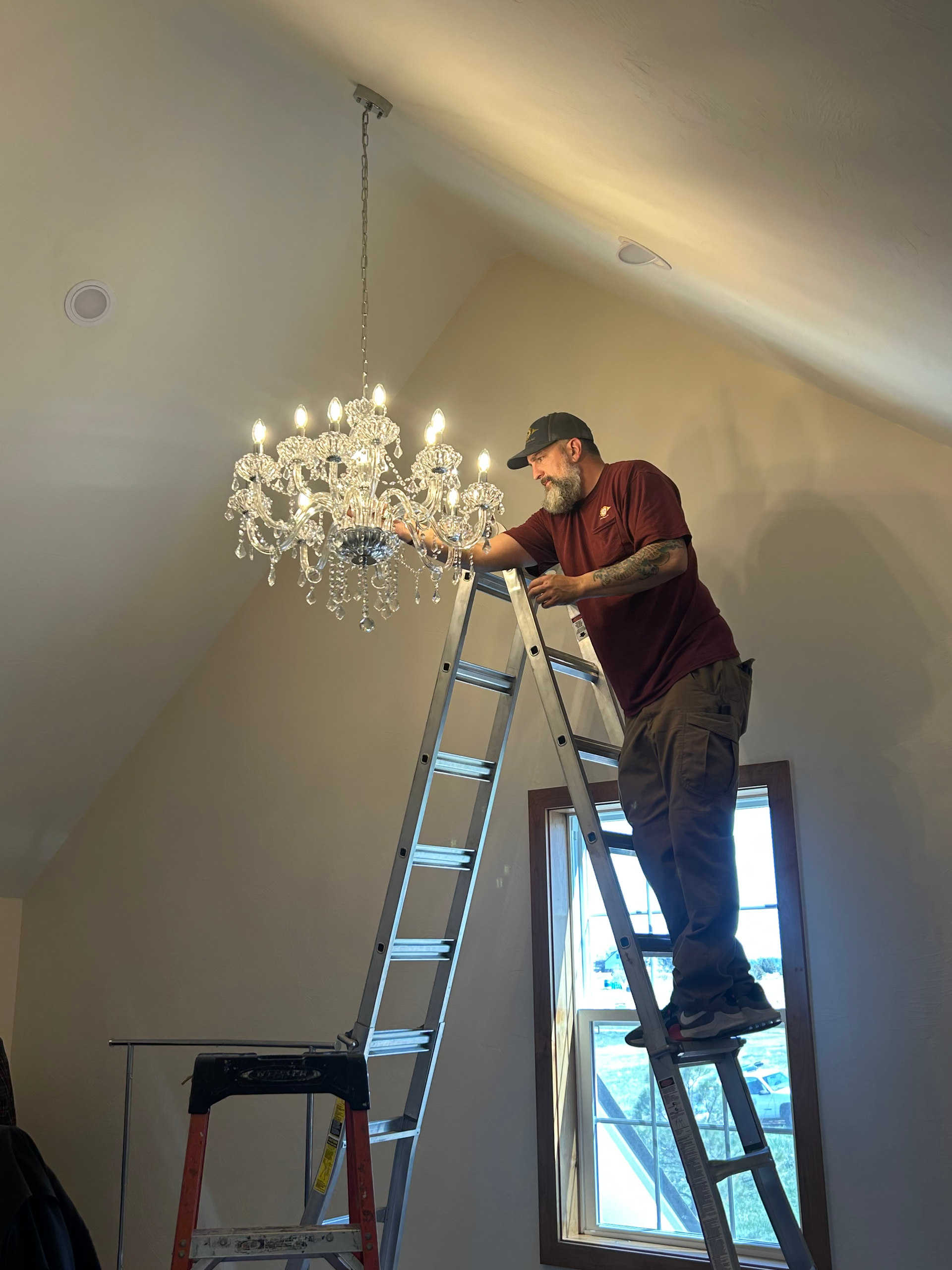 Man on ladder installing a chandelier in a room with a window.