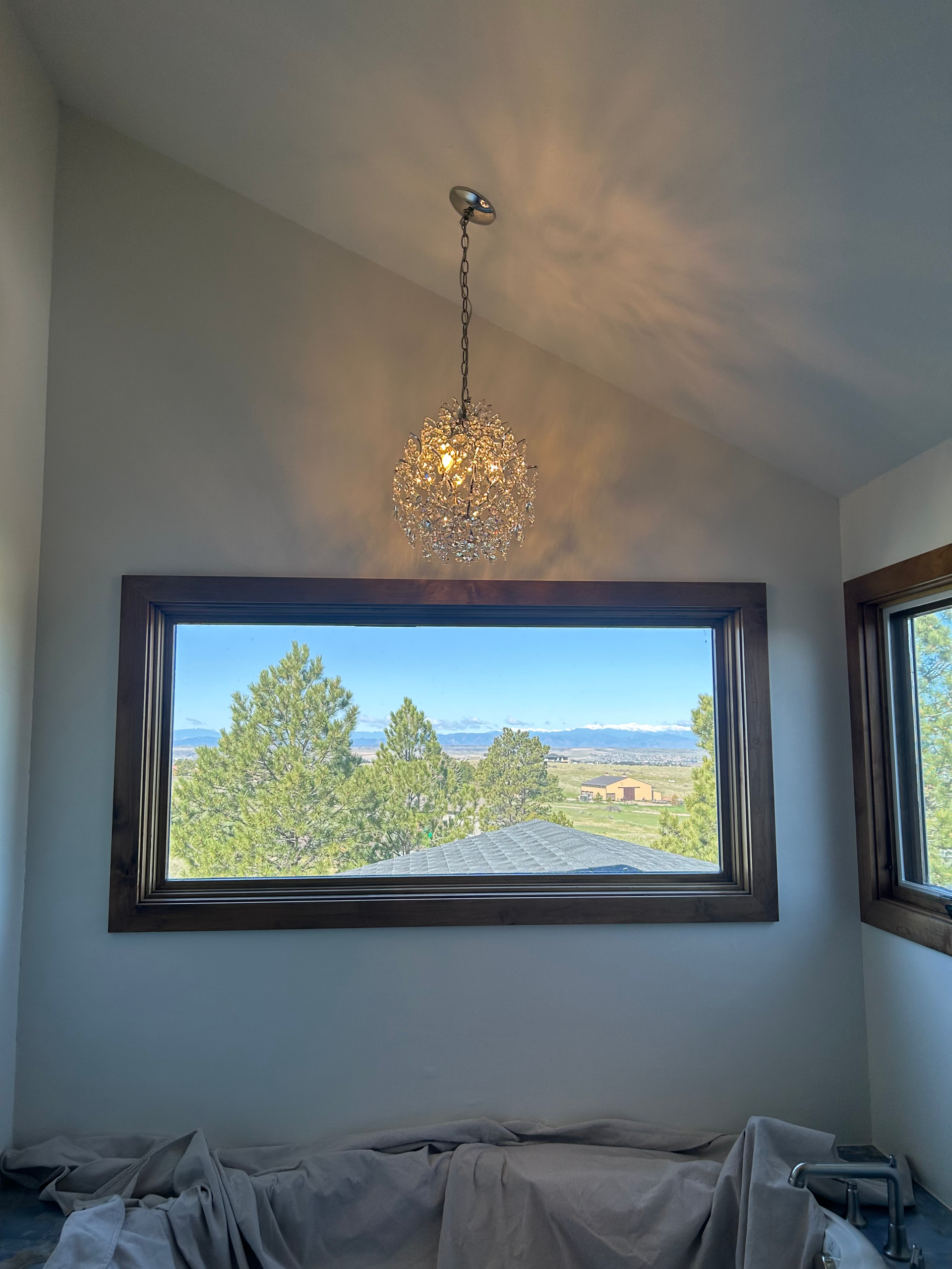 Chandelier hangs above a large window with a scenic view. The room is painted white with dark wooden window frames.