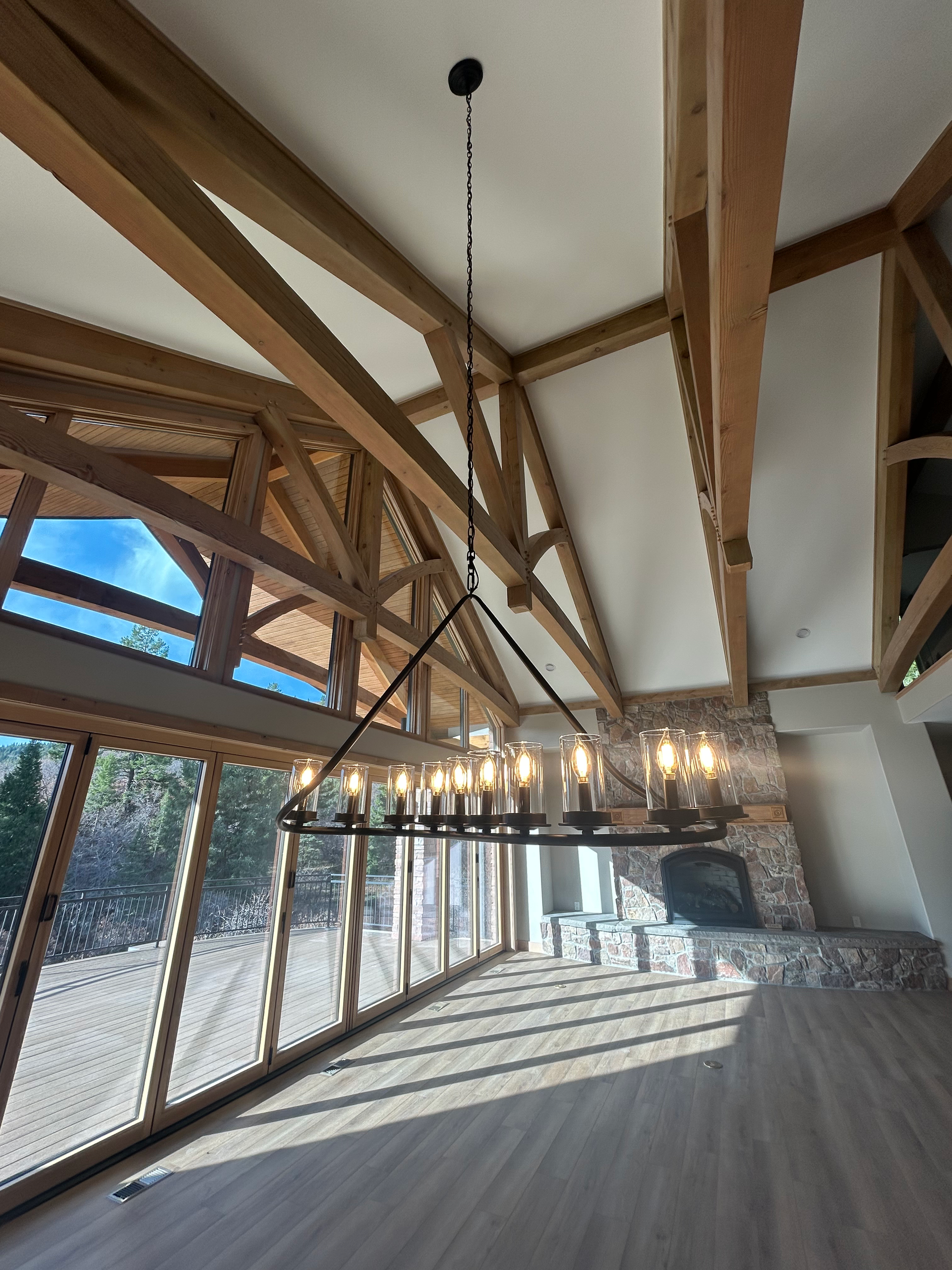 A modern, open-concept living space with exposed wooden beams, large windows, and a black chandelier.
