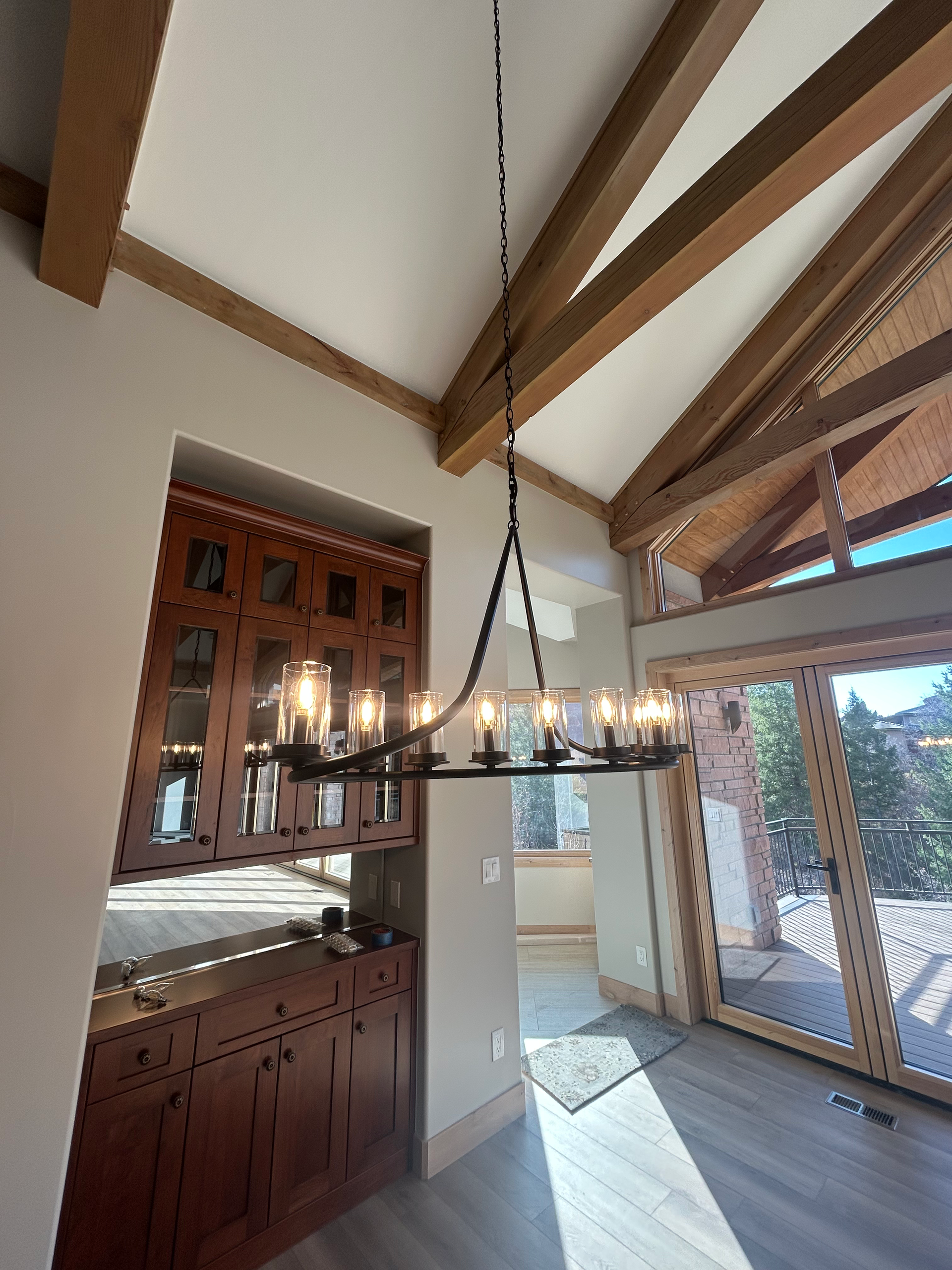 A black chandelier hangs in a room with wood beams, cabinets, and a sliding glass door.